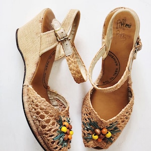 May include: Vintage wedge sandals with woven raffia uppers and straps. The open-toe design features a decorative cluster of orange, yellow, and brown beads with green leaves. The heels are approximately 7.6 cm high.