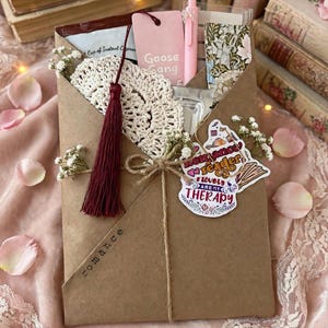 Op de afbeelding: Een bruine papieren envelop met een roze bladwijzer met de tekst Goose Gang, een roze pen, een wit gehaakt kleedje met een donkerrode kwast en een sticker met de tekst Romance readers are my THERAPY. De envelop is vastgebonden met touw en versierd met gipskruid.