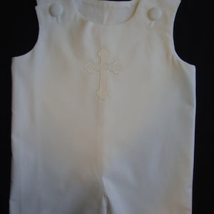 Puede incluir: Romper blanco sin mangas con una cruz bordada en blanco en la parte delantera. El romper tiene dos botones blancos en los hombros.