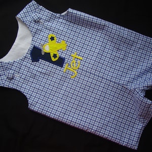 Puede incluir: Un mono de bebé a cuadros azules y blancos con un avión amarillo y el número uno bordado en la parte delantera. El nombre "Jet" también está bordado en el mono.