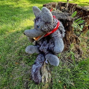 May include: A plush wolf toy with grey, shaggy fur, pointed ears, and a single visible blue eye. The wolf has a wide, open mouth showing a white tooth and wears a bright red bandana patterned with small white shapes around its neck.
