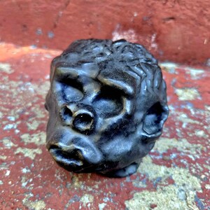 May include: A dark, metallic-toned sculpture of a gorilla head featuring detailed facial features, including a prominent brow, a wide nose, and defined lips. The surface has a textured, weathered appearance with subtle highlights across the contours.