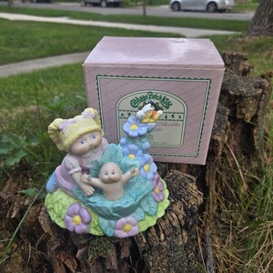 Puede incluir: Figura coleccionable de Cabbage Patch Kids con una niña rubia y un bebé en una cuna con forma de hoja, rodeados de flores. La figura está sobre un tocón de árbol, con la caja original detrás.