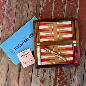 May include: A magnetic backgammon set featuring a wooden board with a cork playing surface, red and white points, white and red checkers, a doubling cube, and a small instruction booklet, all contained in a light blue box labeled BACKGAMMON.