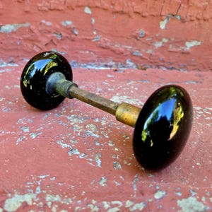 May include: A door knob set featuring two smooth, glossy black spherical knobs connected by a central metallic spindle. The hardware displays a reflective surface on the black knobs and a weathered, tarnished brass finish on the connecting rod.