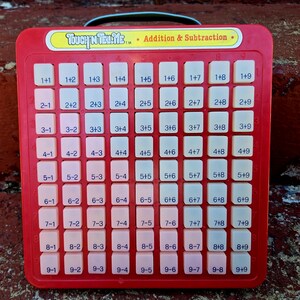 May include: A red plastic Touch 'N Tell Me Addition & Subtraction educational toy featuring a grid of ninety-nine off-white square buttons. Each button is printed with a unique math equation, ranging from 1+1 to 9+9, arranged in rows and columns.