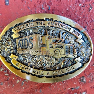 May include: A gold-toned belt buckle with an engraved image of a semi-truck and trailer. The truck has the word "AIDS" on the side and the buckle reads "Truckin' the American Way Every Mile a Safe One".