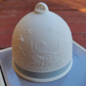 May include: A white ceramic bell-shaped ornament with an embossed winter scene. The design features snowflakes, trees, and a figure. A light blue band encircles the base, and a loop is at the top.