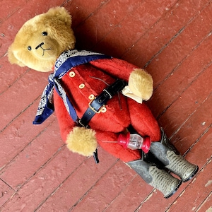 Puede incluir: Un oso de peluche con un atuendo rojo y azul, cinturón negro y botas de vaquero. El oso sostiene un pequeño objeto rojo y blanco en sus patas.