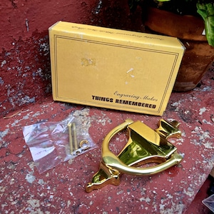 May include: A polished brass door knocker with a triangular base and a curved handle, accompanied by two metal mounting screws and a small washer in a clear plastic bag, presented with a tan cardboard box labeled Engraving Makes THINGS REMEMBERED.