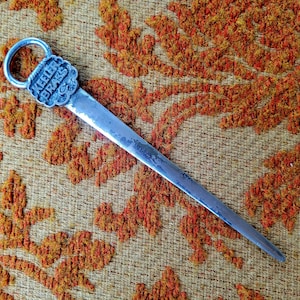 May include: A silver letter opener with a decorative handle featuring the words "Keeler Brass Co.". The letter opener is lying on a patterned orange and brown fabric.