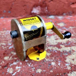 May include: A vintage "Midget" automatic pencil sharpener by the Apsco company. The sharpener is made of metal and has a yellow base and handle. The text "Midget Automatic Pencil Sharpener Company Rockford, ILL., U.S.A." is visible.