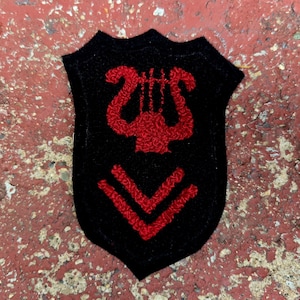 May include: Black shield-shaped patch with red embroidered lyre and chevron design. The patch is likely for a band or orchestra. The background is a textured, red-toned surface.