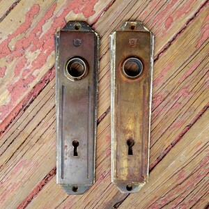 May include: Two antique brass door plates with a keyhole and a round hole for a knob. The plates have a decorative design and are slightly tarnished.