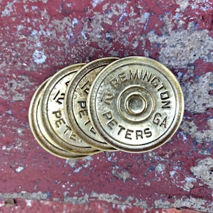 May include: A set of four circular brass shotgun shell heads arranged in a partial stack. Each piece features the embossed text REMINGTON 12 PETERS GA around a central primer pocket. The metallic surfaces have a weathered, aged gold finish.