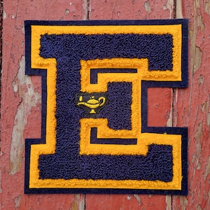 May include: A navy blue felt letter E with a yellow outline. The letter is decorated with a small yellow lamp with a blue flame.