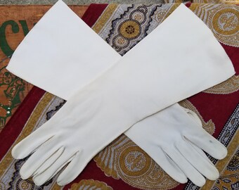 May include: A pair of long white gloves with a smooth texture. The gloves are crossed over each other and are lying on a patterned fabric.