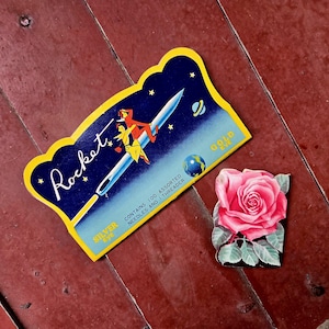 May include: A Rocket brand needle package with a blue background, yellow border, and an illustration of a person riding a rocket, alongside a separate rectangular card featuring a detailed illustration of a single pink rose with green leaves.