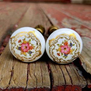 May include: Two white ceramic cabinet knobs with a floral design. Each knob features a pink flower with orange, yellow, and blue accents, framed by gold filigree. The knobs have brass hardware.