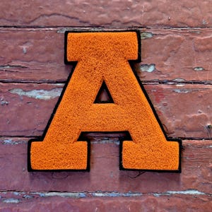 Può includere: Una toppa a forma di lettera A in stile varsity, caratterizzata da una superficie in tessuto ciniglia arancione brillante e testurizzato. La lettera è delineata da un bordo nero spesso e solido che ne definisce la forma a blocchi.