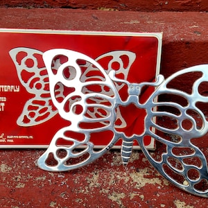 Może przedstawiać: Srebrny, pozłacany stojak na gorące w kształcie motyla w oryginalnym pudełku. Pudełko jest czerwone z białymi napisami: "The Butterfly Trivet, Silver Plated Trivet, Leonard Silverware Company, Inc., Boston, Mass."