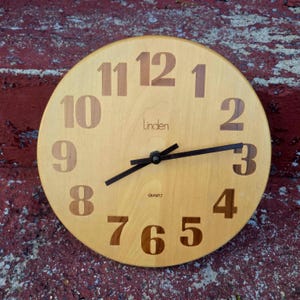 May include: Round wooden wall clock with large, brown numerals and black hands. The clock face is light brown with the brand name "Linden" and the word "QUARTZ" printed on it. The clock is set against a red brick wall.