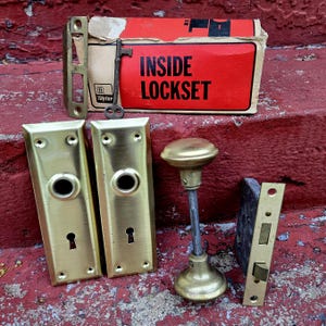 Puede incluir: Un juego de cerradura antiguo con placas de puerta de latón, un pomo y una llave. El juego de cerradura incluye una caja con el texto "INSIDE LOCKSET" en rojo y negro. El hardware se muestra sobre una superficie roja.