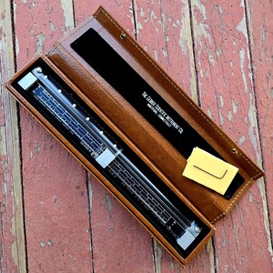 May include: A brown leather case with a slide rule inside. The slide rule is made of metal and glass and has a variety of scales. The case has the text "THE GERBER SCIENTIFIC INSTRUMENT CO. HARTFORD, CONNECTICUT" printed on it.