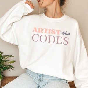May include: White sweatshirt with the text "ARTIST who CODES" in pink and purple lettering.