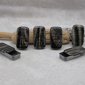 May include: Six small black metal tins with gold glitter lids. The tins are arranged in a row on a wooden surface.