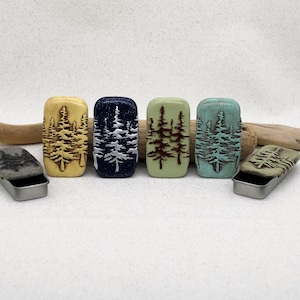 May include: Six rectangular metal tins with lids featuring embossed pine tree designs. The lids come in yellow, dark blue with glitter, light green, turquoise, and two grey versions. Two tins are shown open, revealing their metallic interior.