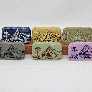 May include: Six rectangular ceramic tiles featuring a relief design of a mountain and a single tree. The tiles are finished in various colours including dark blue, gold, olive green, grey, light green, and light pink, each with a contrasting ink.