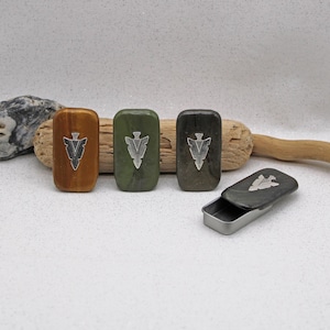 Puede incluir: Cajas rectangulares de madera en tonos marrón, verde y gris, cada una con un diseño de punta de flecha plateada. Una caja está abierta, revelando un interior metálico. Las cajas están dispuestas sobre una superficie blanca con elementos naturales.