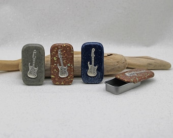 May include: Four small rectangular tins with different colored backgrounds and a silver guitar design. The tins are arranged in a row on a light gray surface. One tin is open and the lid is lying next to it.