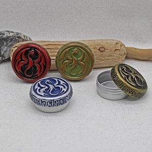 May include: A selection of round containers with dragon designs in red, green, blue, and gold. The lids have embossed dragon motifs. One container is open, showing a silver interior. The containers are suitable for small items.