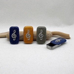May include: Rectangular, colorful trinket boxes with a treble clef design. The boxes are in shades of blue, gold, and gray. One box is open, revealing a silver interior. The boxes are on a white surface with a piece of driftwood.