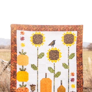 Puede incluir: Un edredón hecho a mano con temática de cosecha otoñal. Muestra girasoles, calabazas y bellotas en tonos amarillos, naranjas y marrones sobre un fondo blanco. El borde es de tela estampada con hojas de otoño.