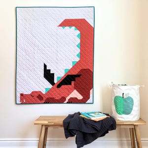 May include: A rectangular quilt featuring a geometric red dragon design on a white background with teal accents and a dark teal border. The dragon has black wings and a long tail. A white fabric storage bin with a green apple print sits nearby.