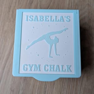 Puede incluir: Un recipiente de tiza de gimnasia azul claro con el texto "ISABELLA'S GYM CHALK". El recipiente presenta un panel blanco con una ilustración de gimnasta. El recipiente de tiza es rectangular.