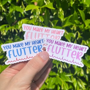 May include: Three white stickers with the text "You make my heart flutter" in pink, blue, and purple lettering. Each sticker has a red, blue, or purple EKG line underneath the text.