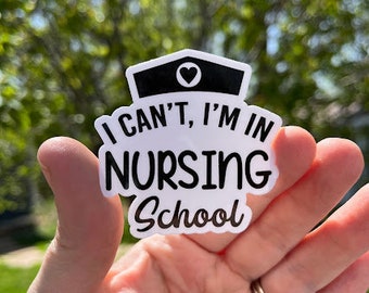May include: A white sticker with black text and a nurse's cap design. The sticker reads "I can't, I'm in Nursing School." The nurse's cap has a black top with a white border and a heart. The sticker is held in a hand.