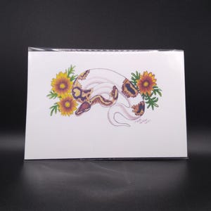 Peut inclure: Une carte blanche avec une illustration à l'aquarelle d'un serpent avec des marques marron et beige, enroulé parmi des tournesols jaunes et orange. La tête du serpent est visible, et les fleurs ont des feuilles vertes. La carte est dans une pochette en plastique transparent.