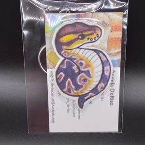 May include: A colorful keychain featuring a snake with a blue and yellow pattern. The keychain is made of wood and has a metal ring for attaching to keys.