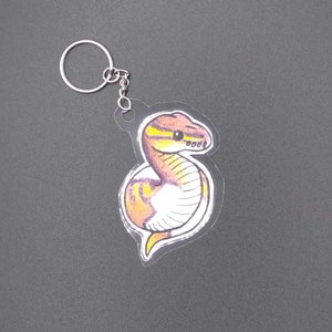 Peut inclure: Un porte-clés en acrylique transparent avec une illustration de serpent jaune, orange et blanc avec des yeux noirs. Le serpent est enroulé en forme de spirale.