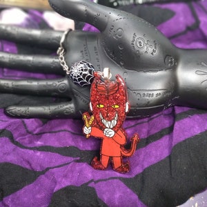 May include: A red glitter devil charm with a slingshot and a toothy grin. The charm is hanging from a silver chain with a black spider web charm.