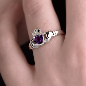 Peut inclure: Une bague Claddagh en argent avec une pierre précieuse en forme de cœur violette. La bague est portée à un doigt.