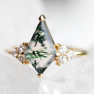 Peut inclure: Une bague en or avec une pierre centrale en agate mousse en forme de cerf-volant présentant des inclusions vertes. La pierre est flanquée de chaque côté par un groupe de trois petites pierres blanches rondes serties sur une fine bague en or.