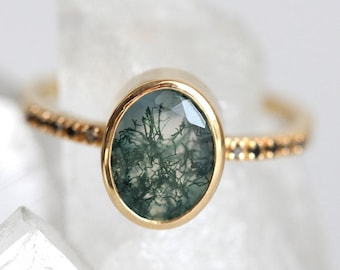 May include: A gold ring featuring an oval-shaped moss agate gemstone with dark green, branch-like inclusions. The band is thin and metallic, accented with small, dark, evenly spaced stones along the sides of the ring shank.