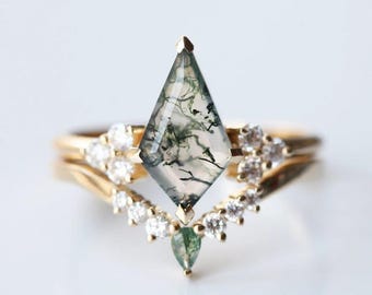 Può includere: Un anello in oro con una pietra centrale in agata muschiata a forma di aquilone con inclusioni verde scuro. La fascia è decorata con piccole pietre bianche rotonde a chevron, con una piccola agata muschiata a goccia alla base.