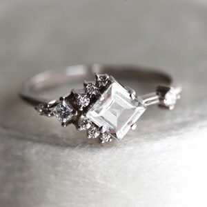Peut inclure: Une bague en argent avec une grande pierre précieuse carrée transparente entourée de petites pierres précieuses scintillantes.